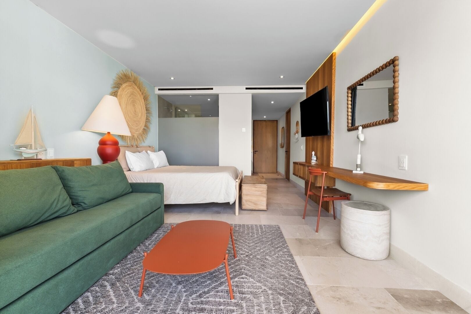 Quarto de hotel moderno com um sofá verde, uma cama, uma mesinha, uma escrivaninha, uma TV e um espelho.