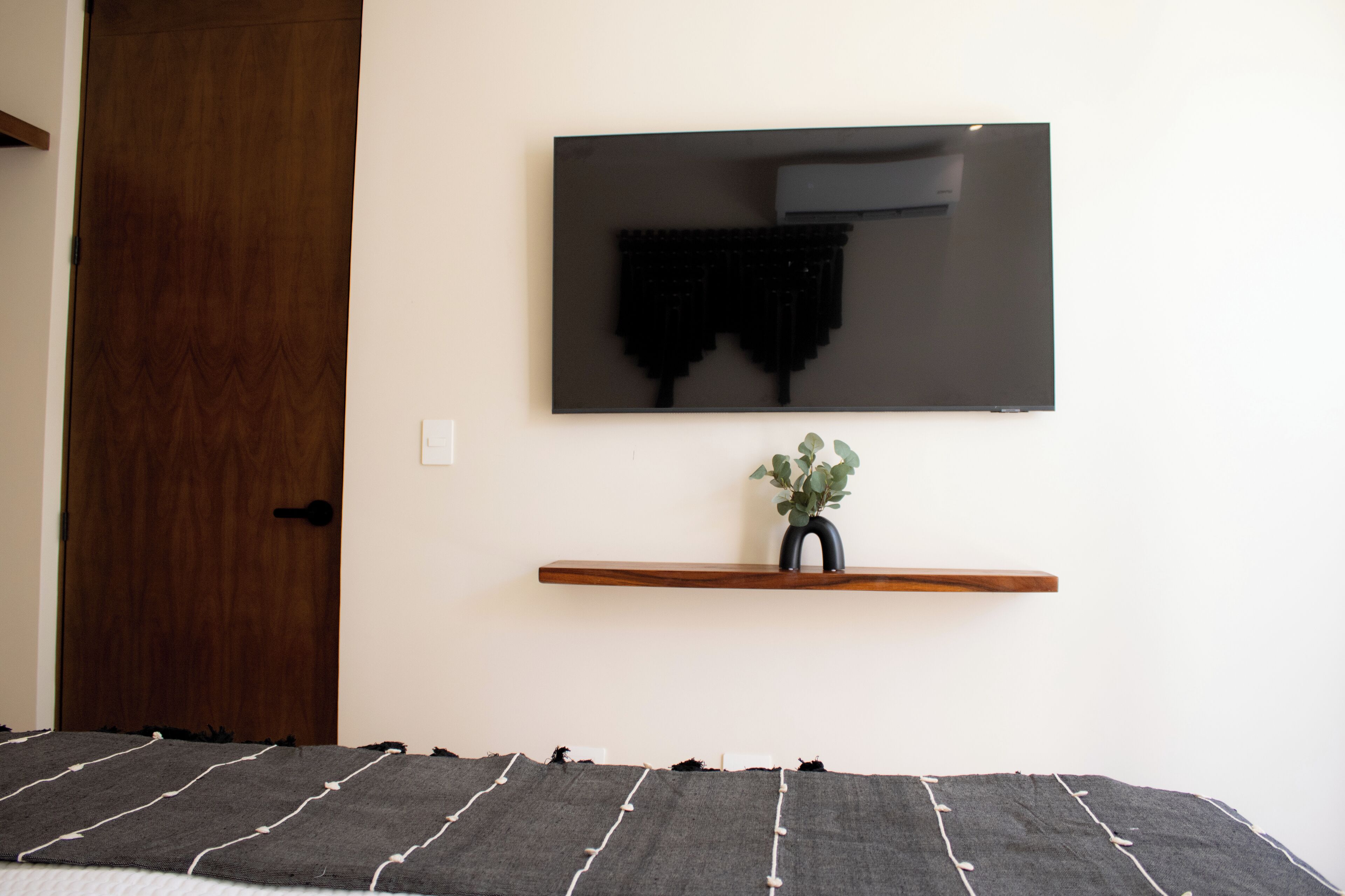 Une chambre avec une porte en bois, un lit recouvert d’une couverture à rayures, un téléviseur fixé au mur et une petite étagère avec une plante.