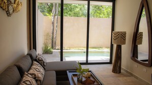A modern living room with a sofa, a tray with decorative items, and a large window offering a view of a pool and greenery. - Peregrina Tulum 101A 2BR Luxury Condo (Tulum)