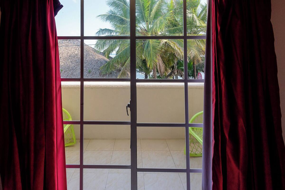 A room with a sliding glass door leading to a balcony with a tiled floor and a view of palm trees and a mountain.