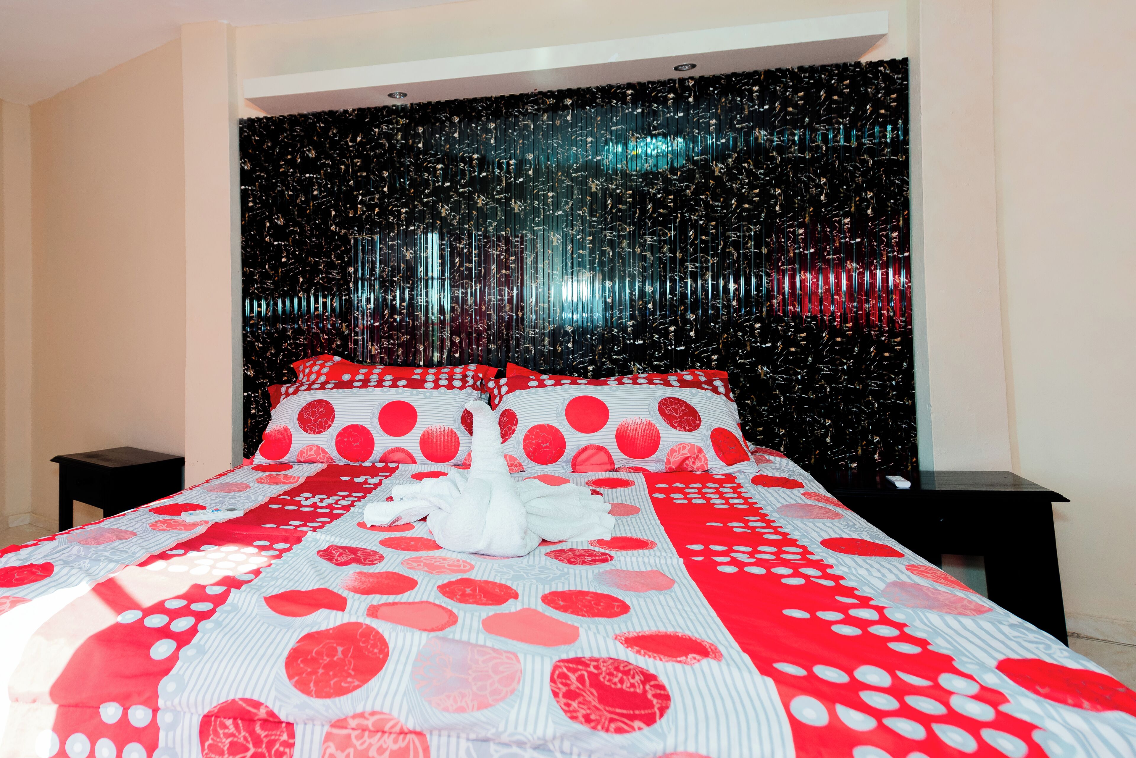 Uma cama com colcha estampada em vermelho e branco, duas mesinhas de cabeceira pretas e uma cabeceira preta com superfície refletora.