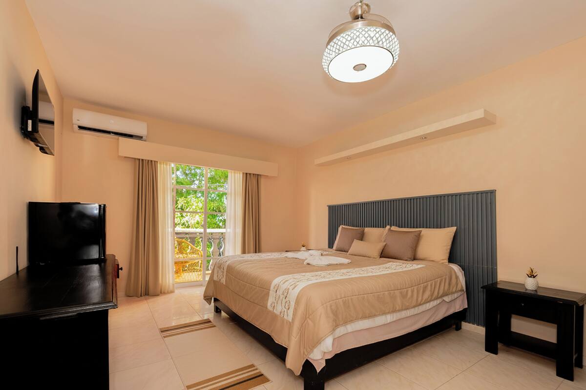 A bedroom with a bed, a TV, an air conditioner, a ceiling light, and a window with curtains.