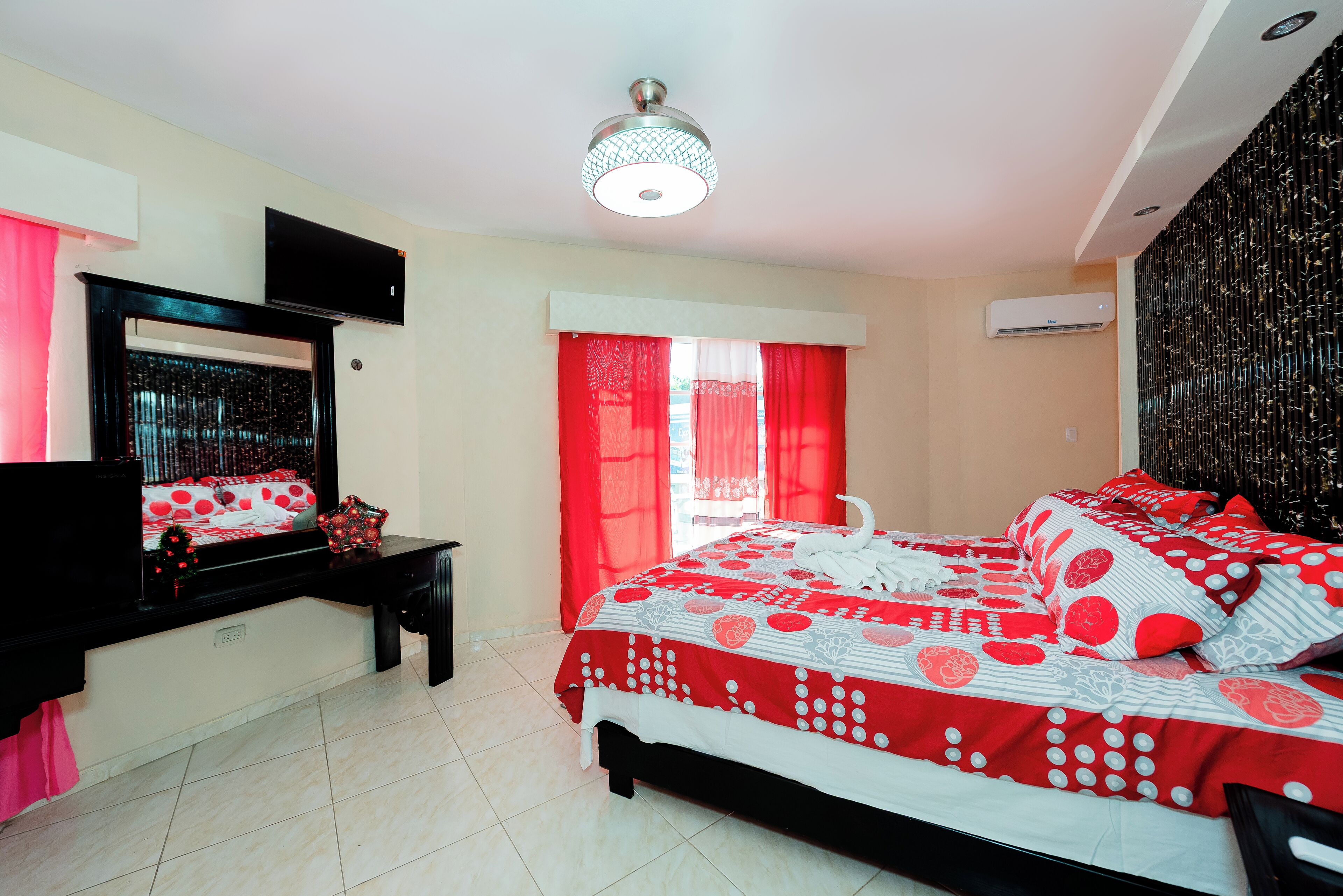 A bedroom with a bed, a TV, a window with red curtains, and a ceiling light.