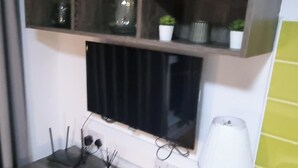 Una habitación con un televisor montado en la pared, una lámpara sobre una mesita auxiliar y una estantería encima del televisor con adornos.