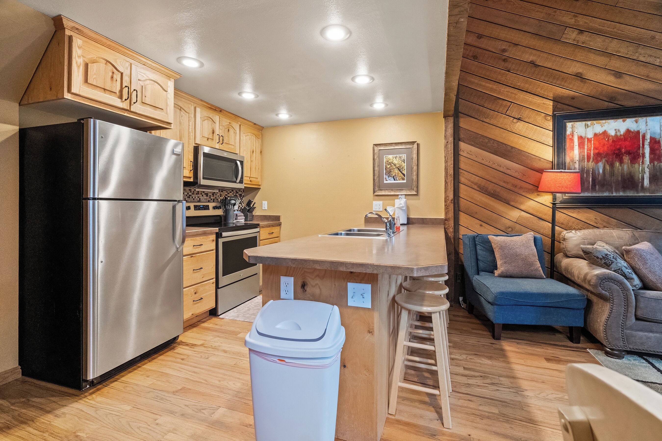A compact kitchen with wooden cabinets, a stainless steel refrigerator, a microwave, and a sink. There is a wooden dining table with a chair and a blue sofa in the adjacent living area.