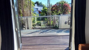 A room with a glass door leading to a balcony with a wooden deck, a view of trees and a house in the background.