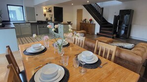 A dining area with a wooden table set for four, surrounded by leather chairs. A kitchen with modern appliances and a staircase leading to an upper level is visible in the background.