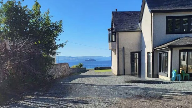 A building with a gravel driveway leading to a door, a view of the sea, and a tree to the left.