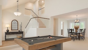 Un vaste espace de vie comprenant une table de billard, une table à manger avec des chaises, un escalier et un lustre.