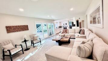 A spacious living room with a large beige sofa, a coffee table, and two armchairs. There is a kitchen area in the background with white cabinets and a microwave.