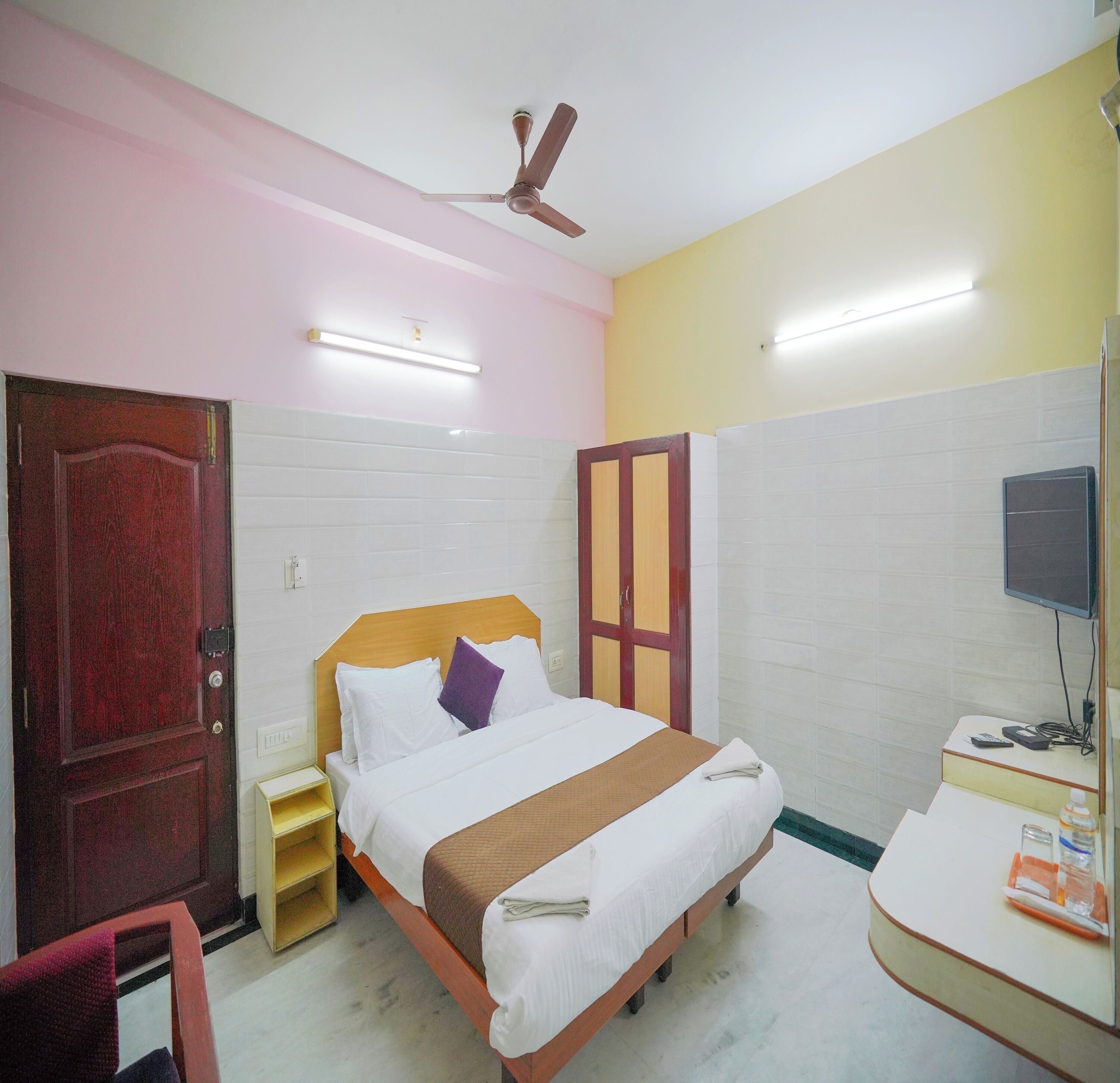 A hotel room with a bed, a desk, a TV, a ceiling fan, and a door.
