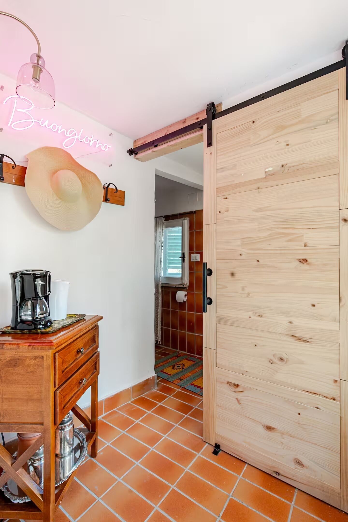 Una habitación con puerta corredera de madera, una cafetera sobre un mueble de madera, un letrero de neón y una alfombra estampada.