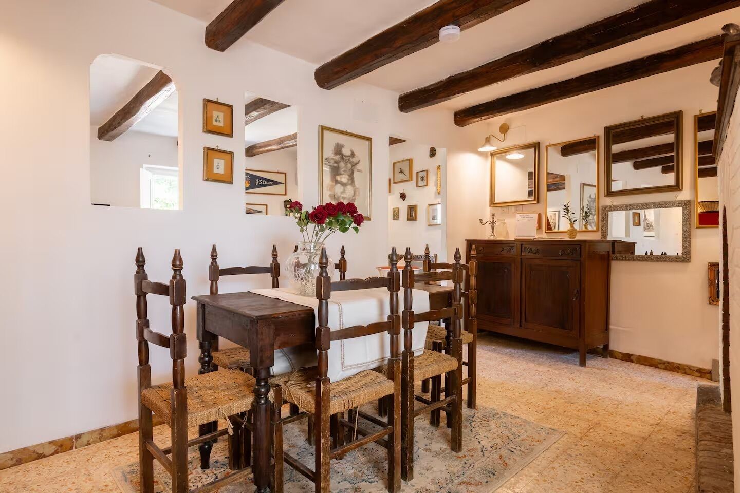 A dining area with a wooden table and chairs, a vase with red flowers, and a cabinet with mirrors.