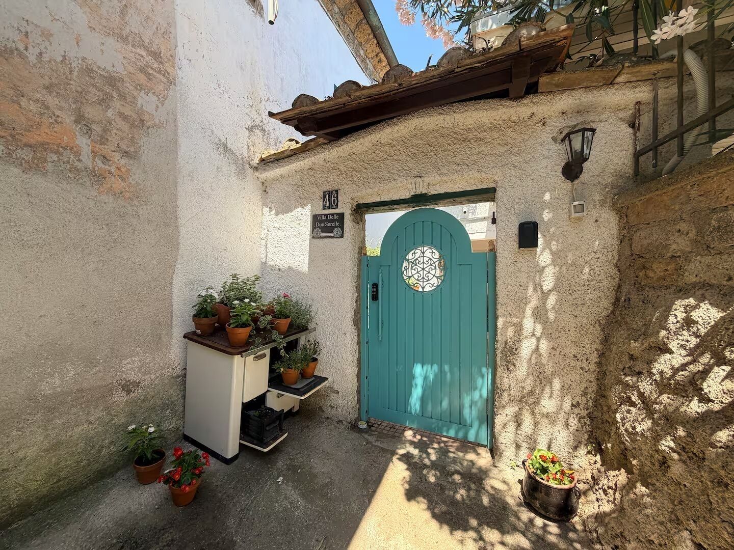 Un vicolo stretto con una porta color petrolio, piante in vaso e un piccolo armadietto bianco.