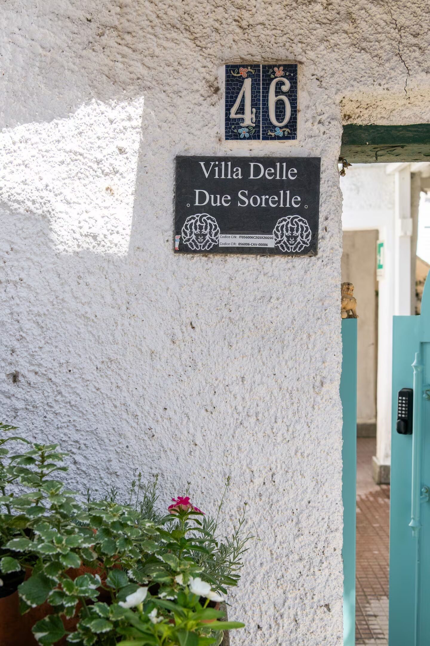 A house number sign, a villa name sign, and a small statue on a textured wall.