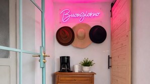 A narrow hallway with terracotta tiles, a wooden table with a coffee maker and a plant, and a neon sign reading "Buongiorno".