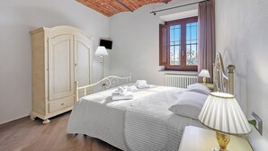 A bedroom with a brick ceiling, a wooden wardrobe, a bed with white bedding, a nightstand with a lamp, and a window with curtains.