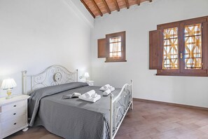 Un lit bien fait, recouvert d’une couvre-lit grise, une table de chevet avec une lampe, et deux fenêtres dotées de volets en bois.