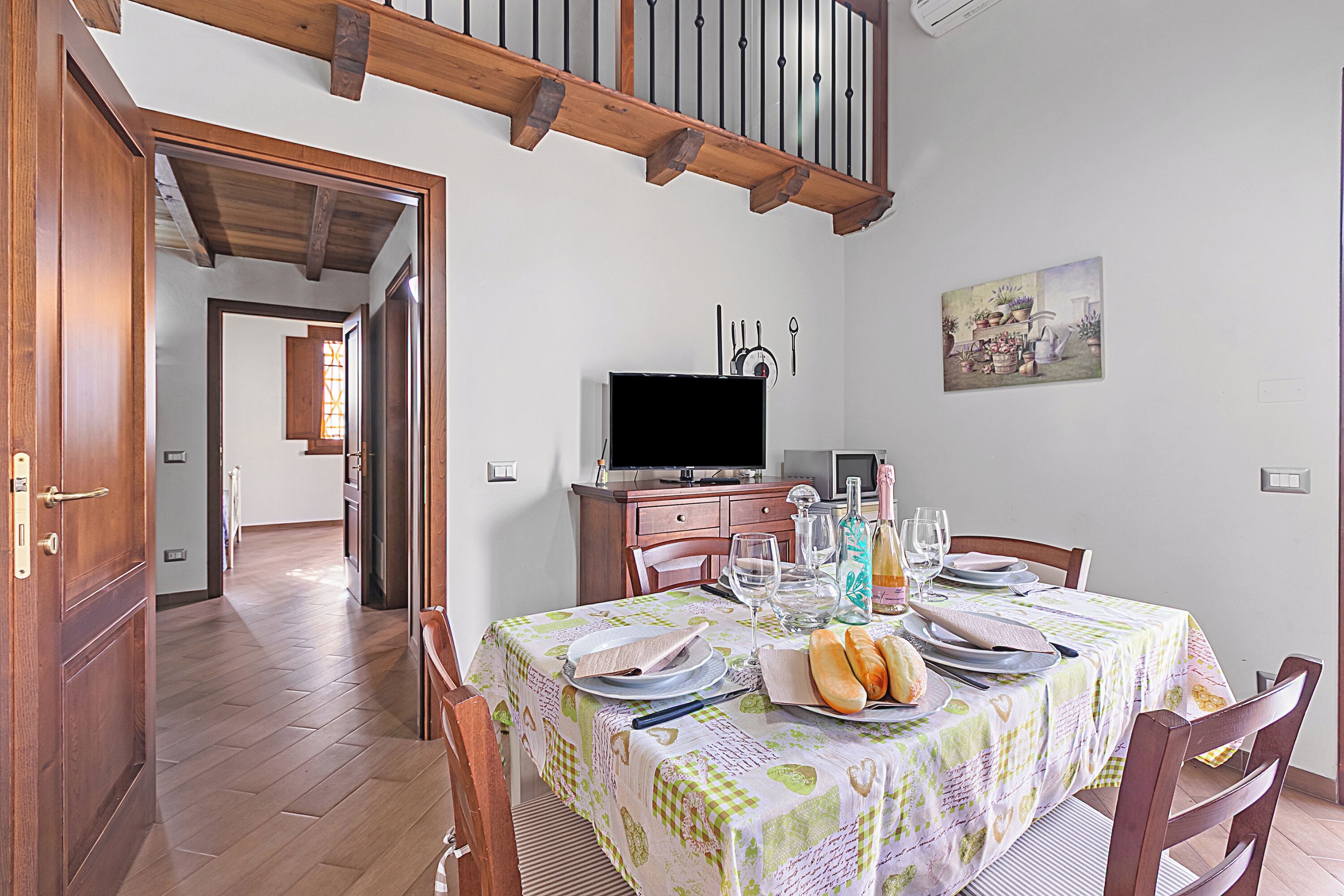 A dining area with a table set for four, wooden chairs, a television on a stand, and a painting on the wall.