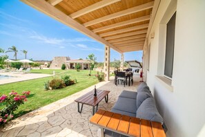 A covered outdoor patio with a stone floor, a wooden table, a sofa, and a view of a swimming pool and a stone building in the background. - Trullo Tenuta Mary with Heated Pool, Carpignano Salentino, Italy (Carpignano Salentino)