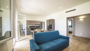 A modern living room with a blue sofa, a dining area with a table and chairs, and a kitchen in the background. - Trullo Tenuta Mary with Heated Pool, Carpignano Salentino, Italy (Carpignano Salentino)