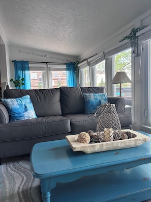 Un salon avec un canapé gris, des coussins bleus, une table basse en bois agrémentée d’objets décoratifs et un mur orné d’une citation.