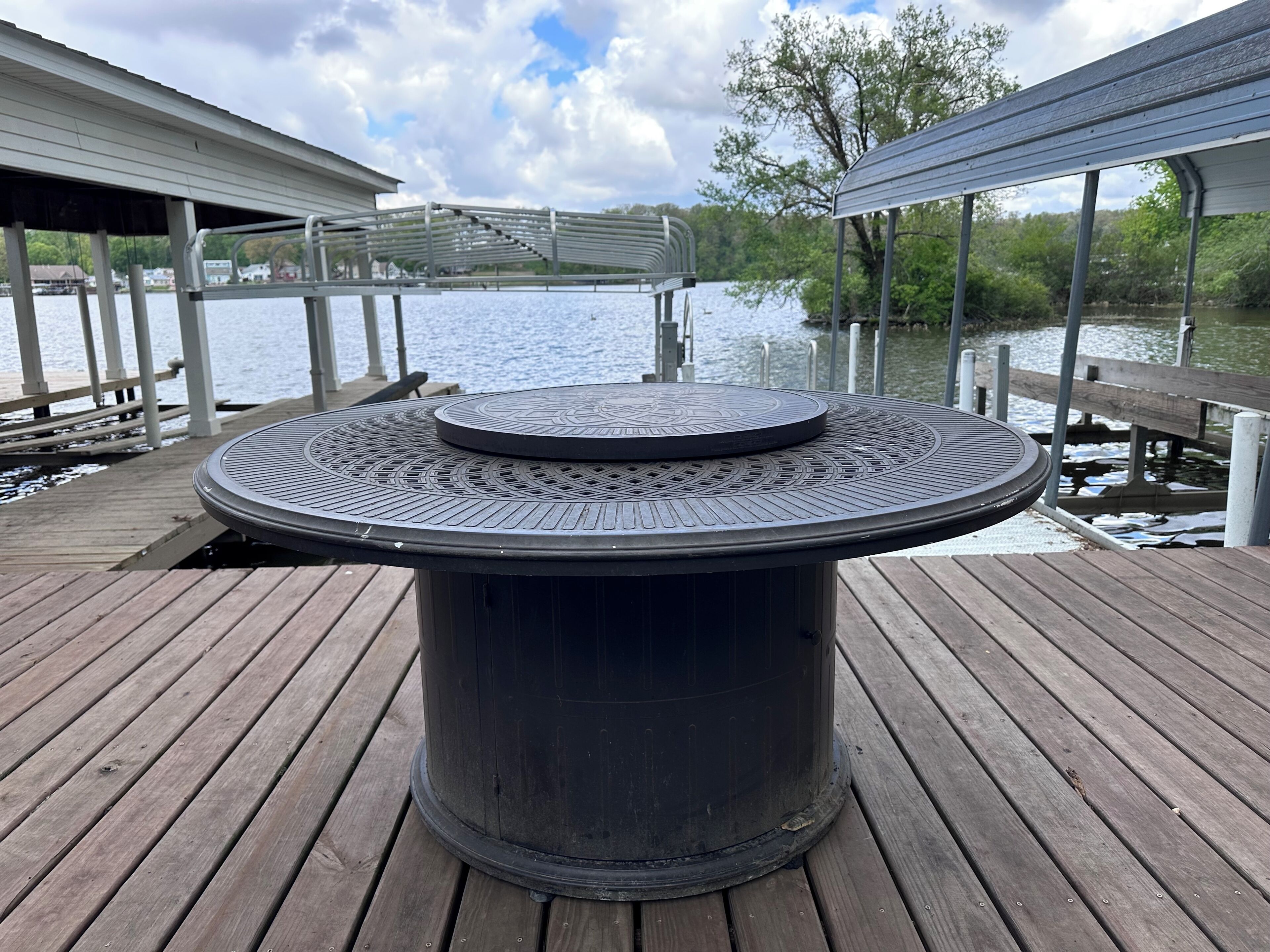 A round outdoor table with a metal top, located on a wooden deck with a view of a lake and dock structures.