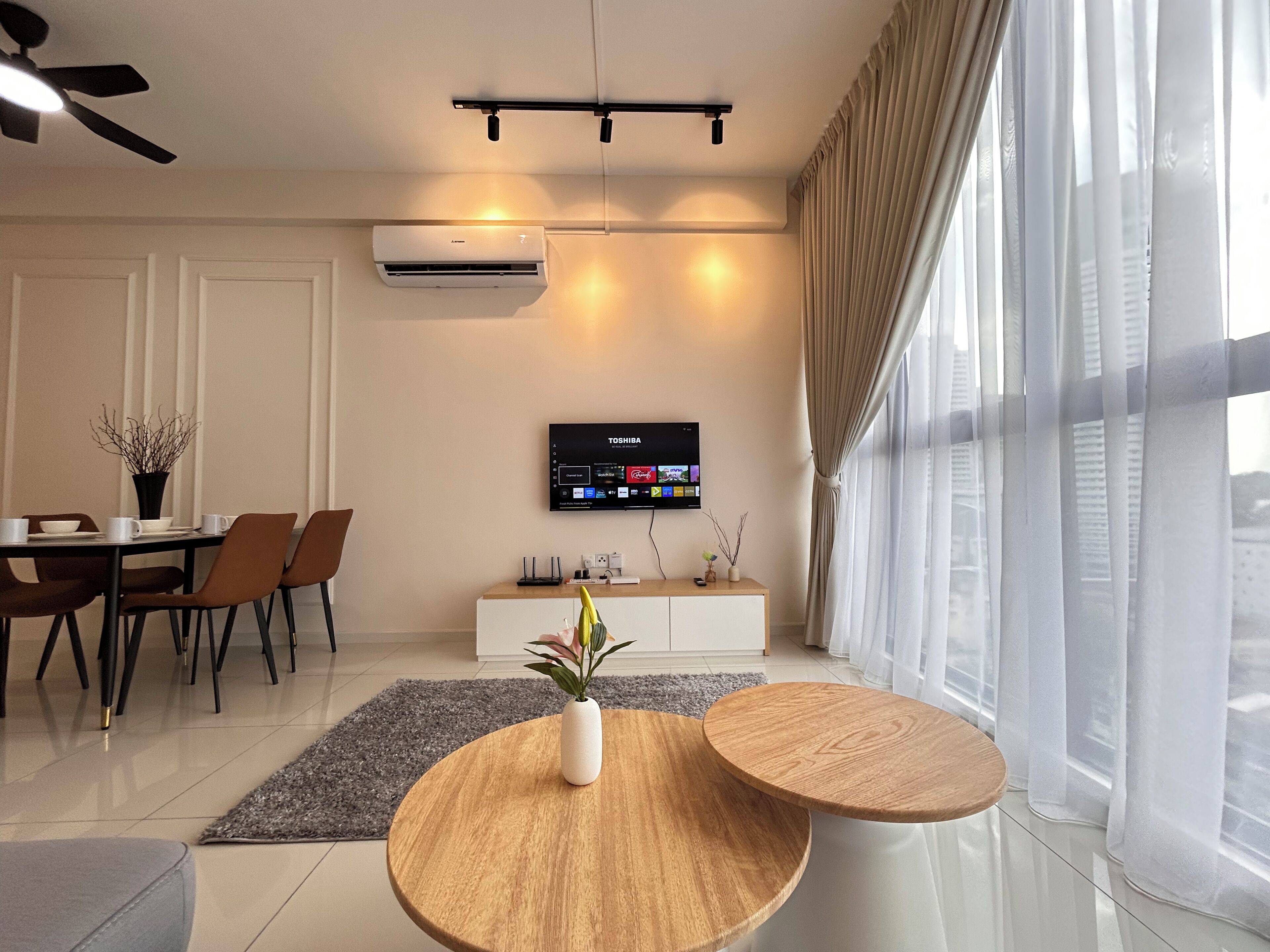Una sala de estar moderna con comedor, un televisor de pantalla plana en la pared y un amplio ventanal con cortinas translúcidas.