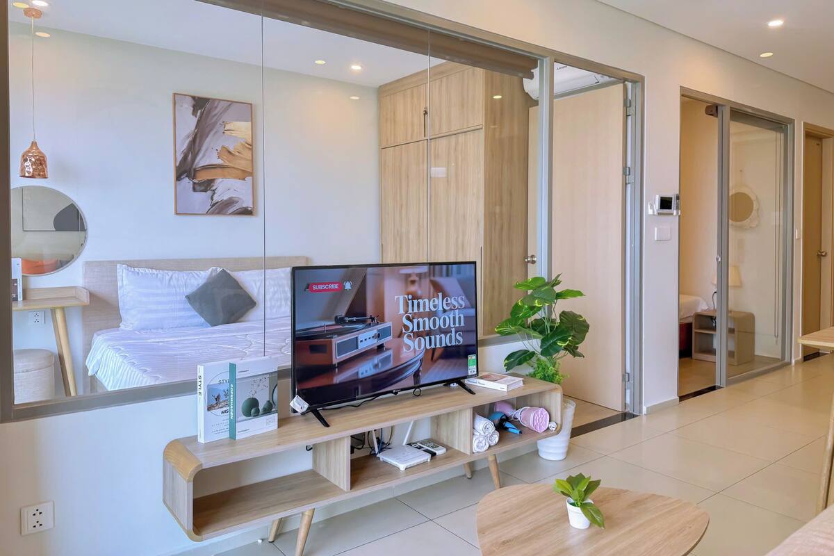 A modern hotel room with a large mirror, a flat-screen TV, a wooden TV stand, a bed, a small table, and a potted plant.