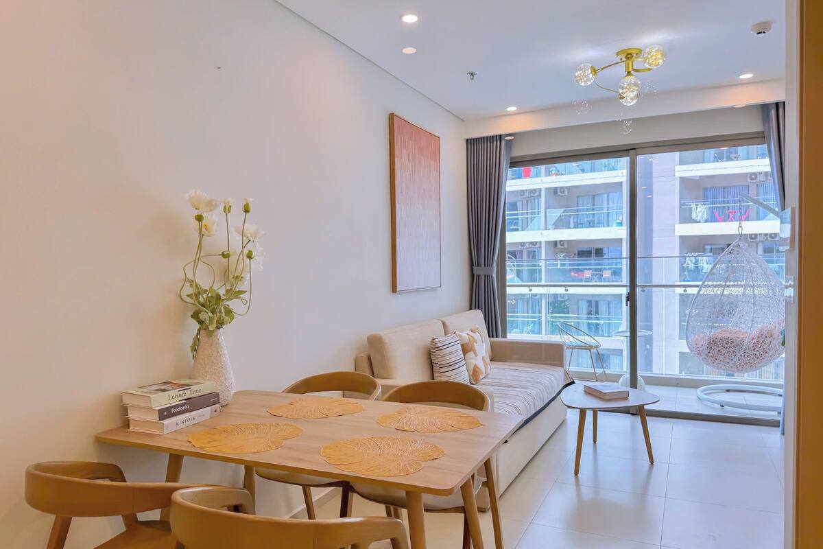 A modern living room with a sofa, dining table, and chairs. There is a vase with flowers on the table, and a hanging chair near the window.