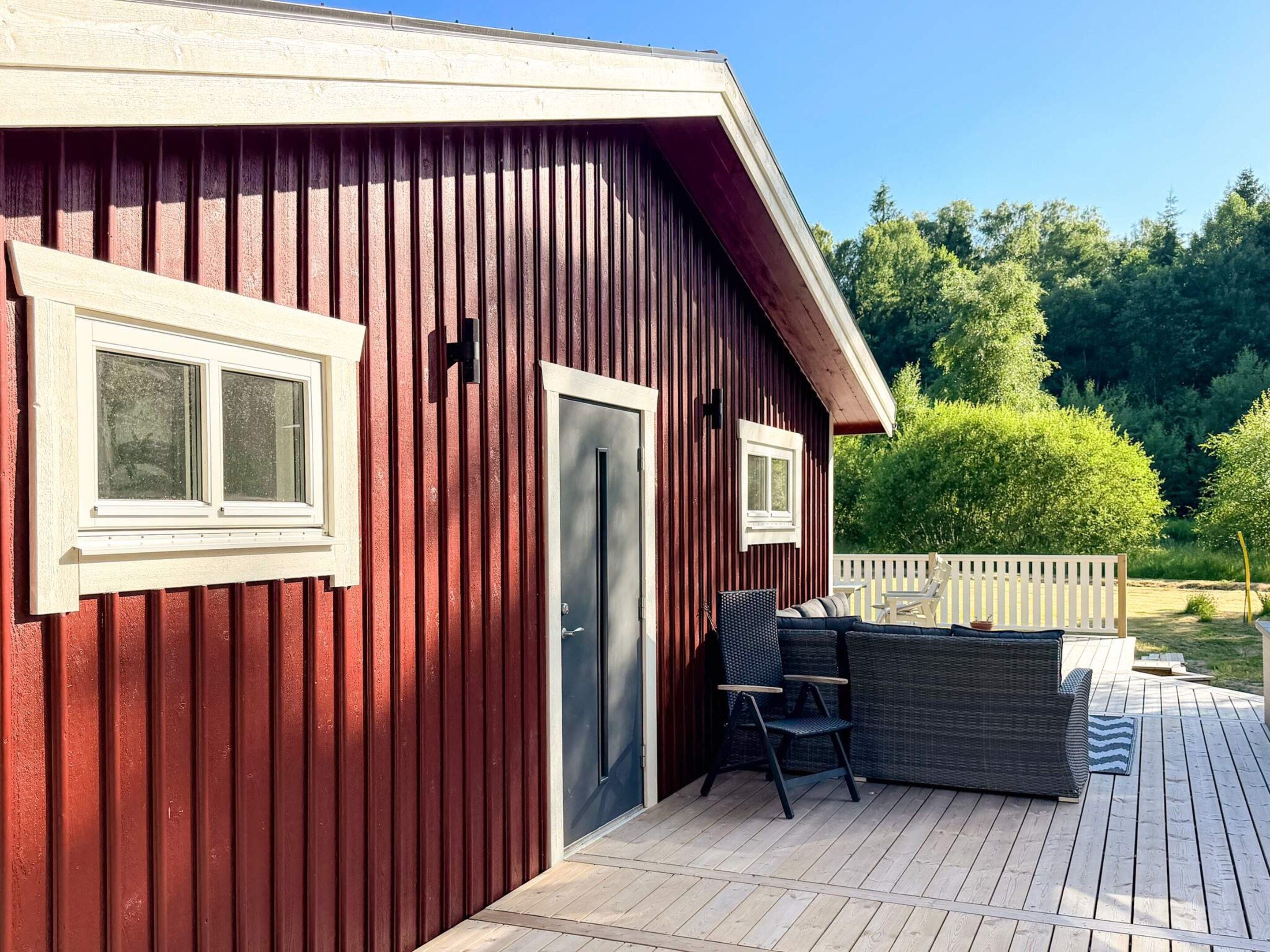 A red wooden building with a door and windows, a deck with outdoor furniture, and a white picket fence in the background.