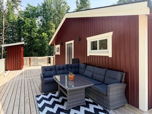 A covered outdoor seating area with a dark-colored sofa and a glass-top table, a wooden deck, and a red building with a door and windows. - 6 Person Holiday Home in Henan (Henån)