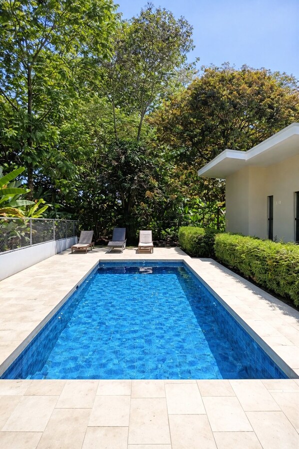Una piscina rectangular con bordes de azulejos, rodeada por una terraza también de azulejos. Hay dos sillas reclinables y una pequeña mesa.