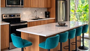 Een moderne keuken met houten kastjes, roestvrijstalen apparatuur, een marmeren aanrecht en teal barstoelen.