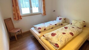 Une petite chambre avec un lit simple, une chaise en bois, une fenêtre avec des rideaux et un mur uni.