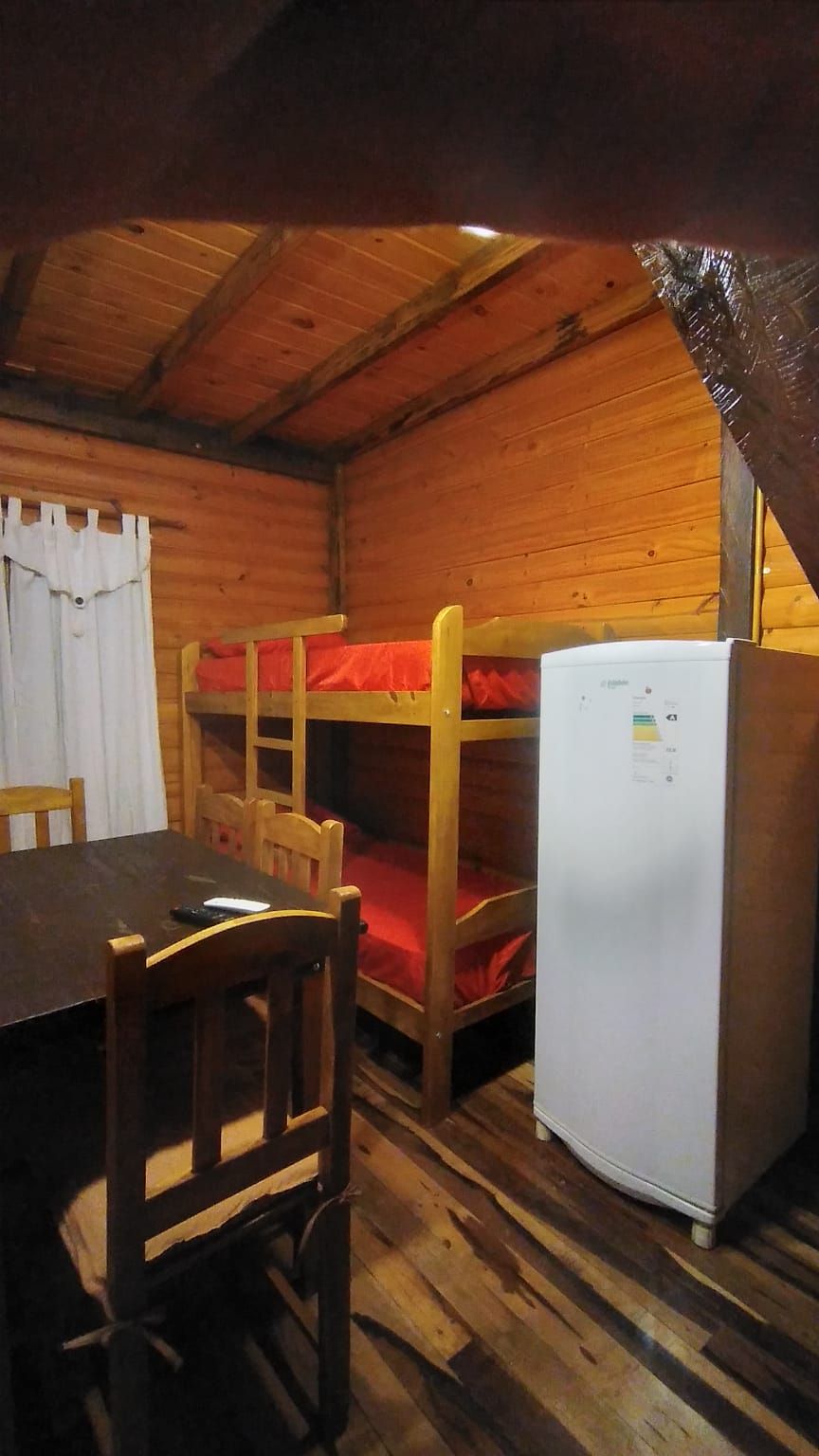 A bunk bed room with a wooden table and chairs, a refrigerator, and a window with curtains.