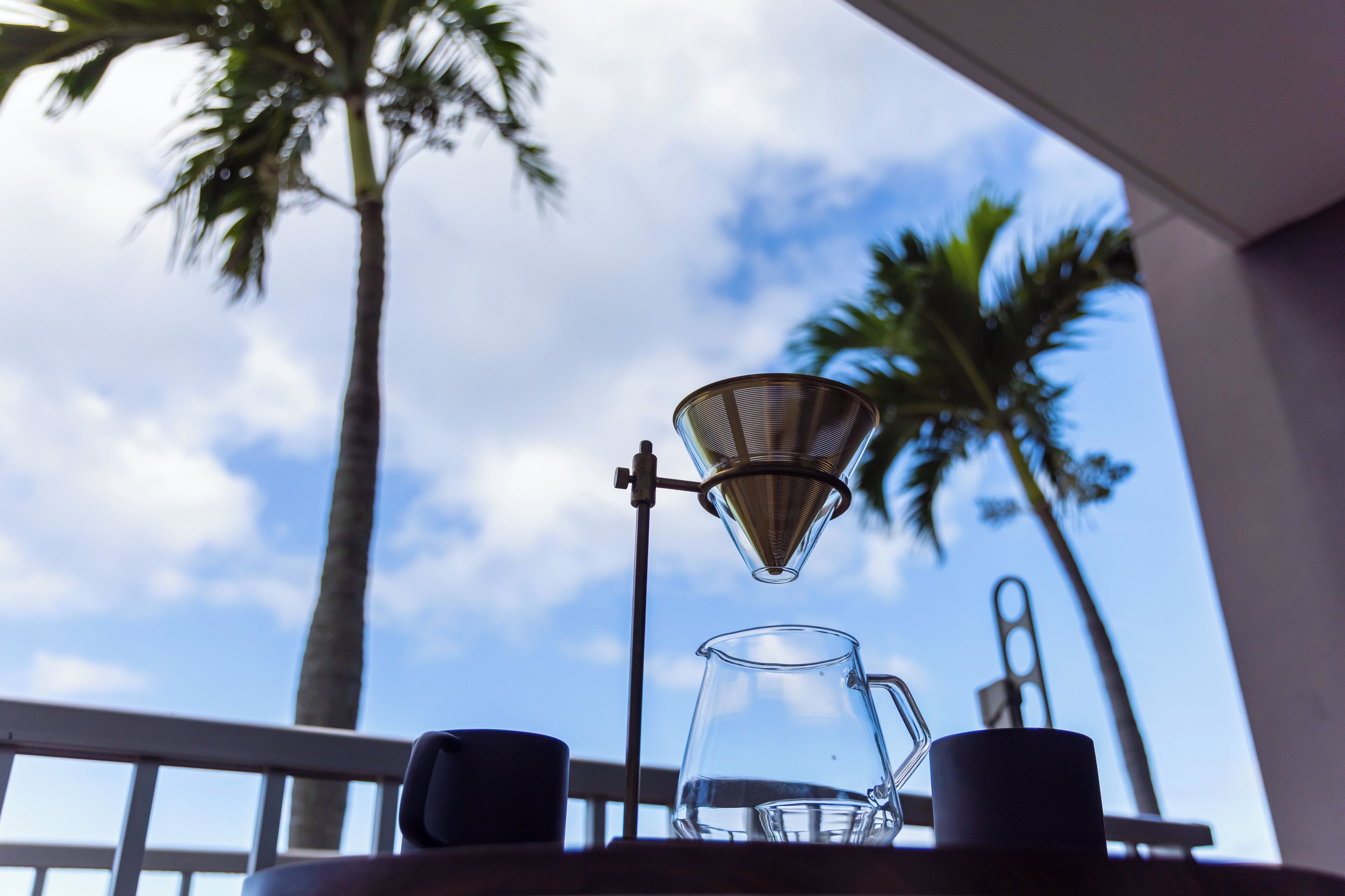 A coffee maker on a balcony with a clear view of palm trees and a blue sky.