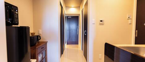 A narrow hallway with a kitchen area on the left, featuring a refrigerator, microwave, and coffee maker. The hallway has a tiled floor and a ceiling with recessed lighting.