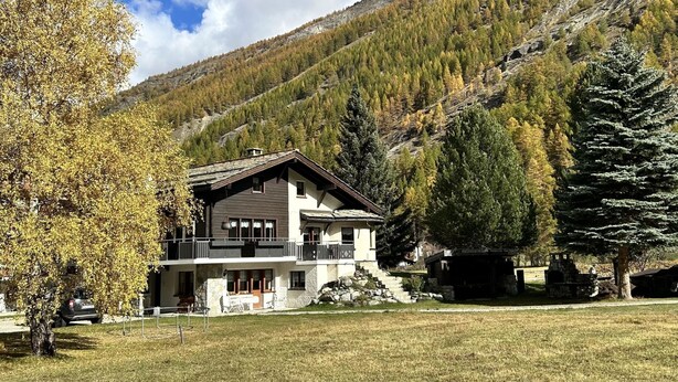 Un chalet de dos pisos con balcón, rodeado de césped y árboles, con montañas al fondo.