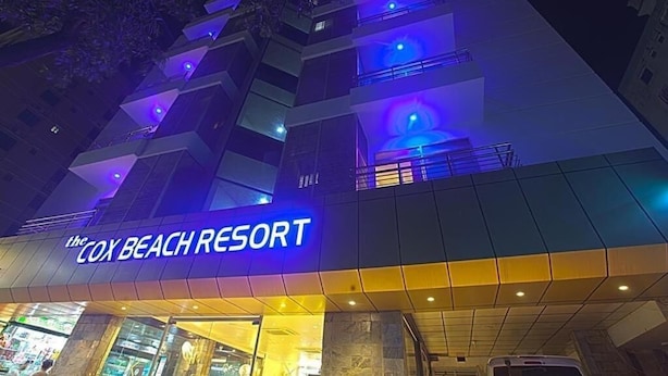 La façade du bâtiment du complexe hôtelier Cox Beach Resort, de nuit, avec ses enseignes illuminées et un éclairage bleu.