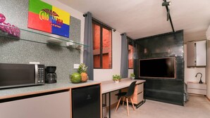 A compact kitchen with a microwave, coffee maker, and a small refrigerator. There is a dining area with a chair and a table, and a large TV mounted on a textured wall. - 1BR-1AC Stylish Studio near Provenza&Parque Lleras (Medellín)
