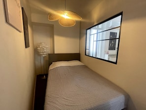 Une chambre d’hôtel étroite, avec un lit simple, une grande fenêtre et un luminaire suspendu.