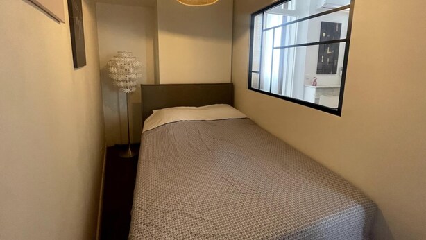 Ein schmales Hotelzimmer mit einem Bett, einem großen Fenster, einer Stehlampe und einer Wandleuchte.