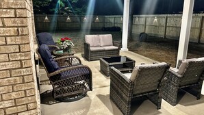 An outdoor patio area with wicker furniture, including chairs and a sofa, under a covered structure with a view of a wooden fence and trees in the background.