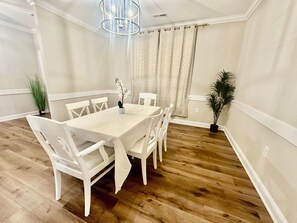 Uma sala de jantar com uma mesa retangular, cadeiras brancas, um candelabro e um vaso com planta.