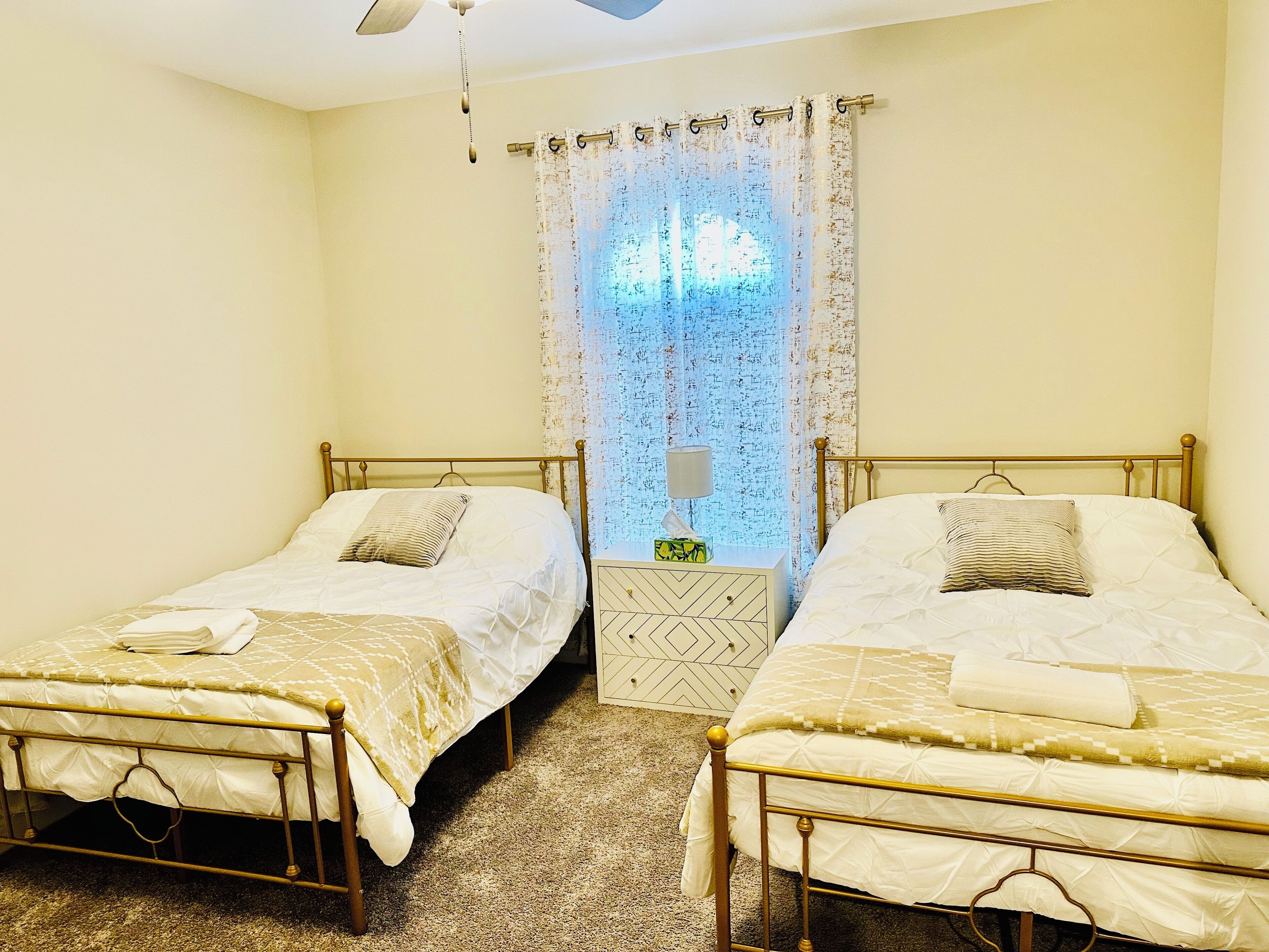 Habitación doble con dos camas individuales, una mesita de noche, una lámpara y una ventana con cortinas estampadas.