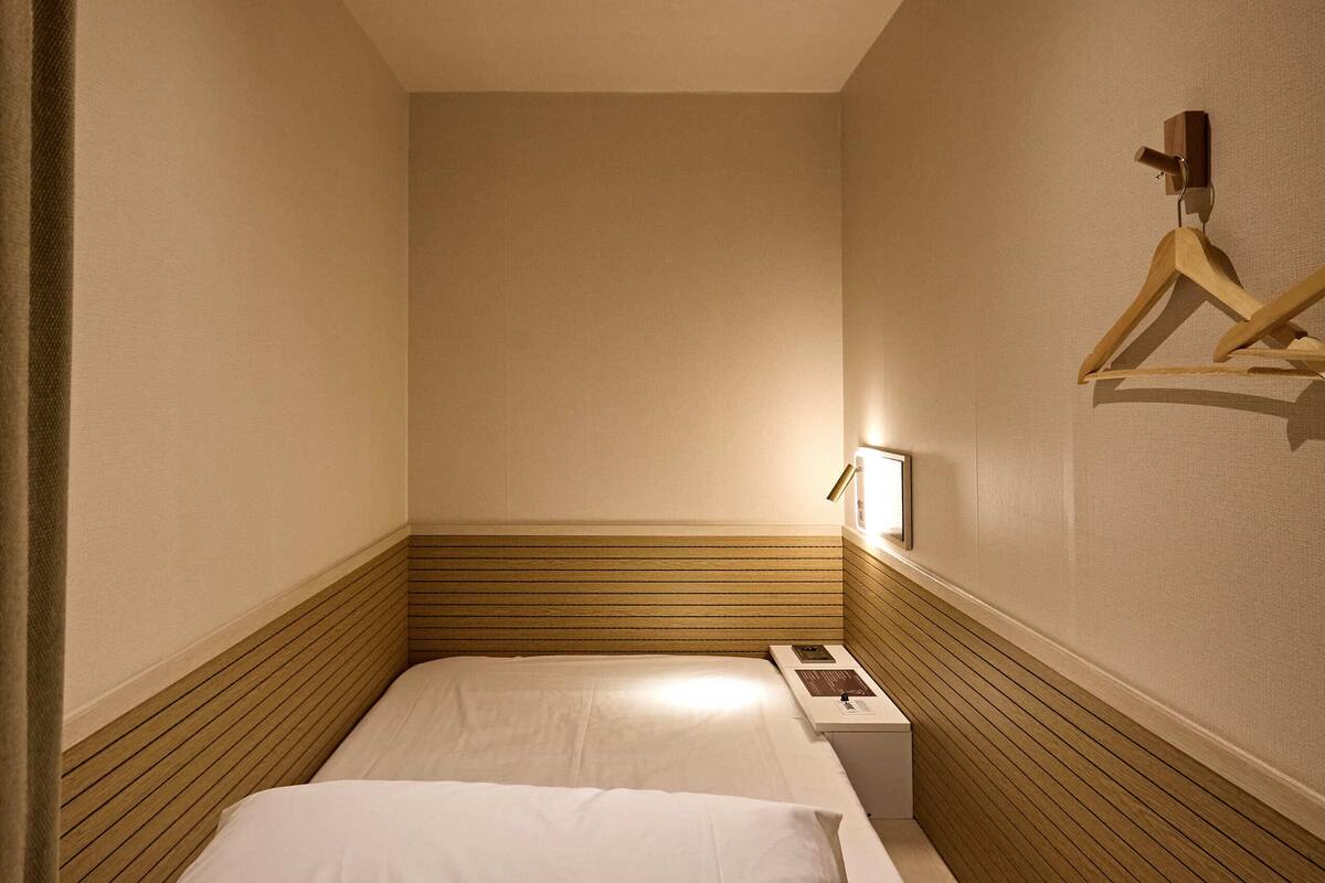 A narrow hotel room with two beds, a bedside table, a hanging coat rack, and a ceiling light.