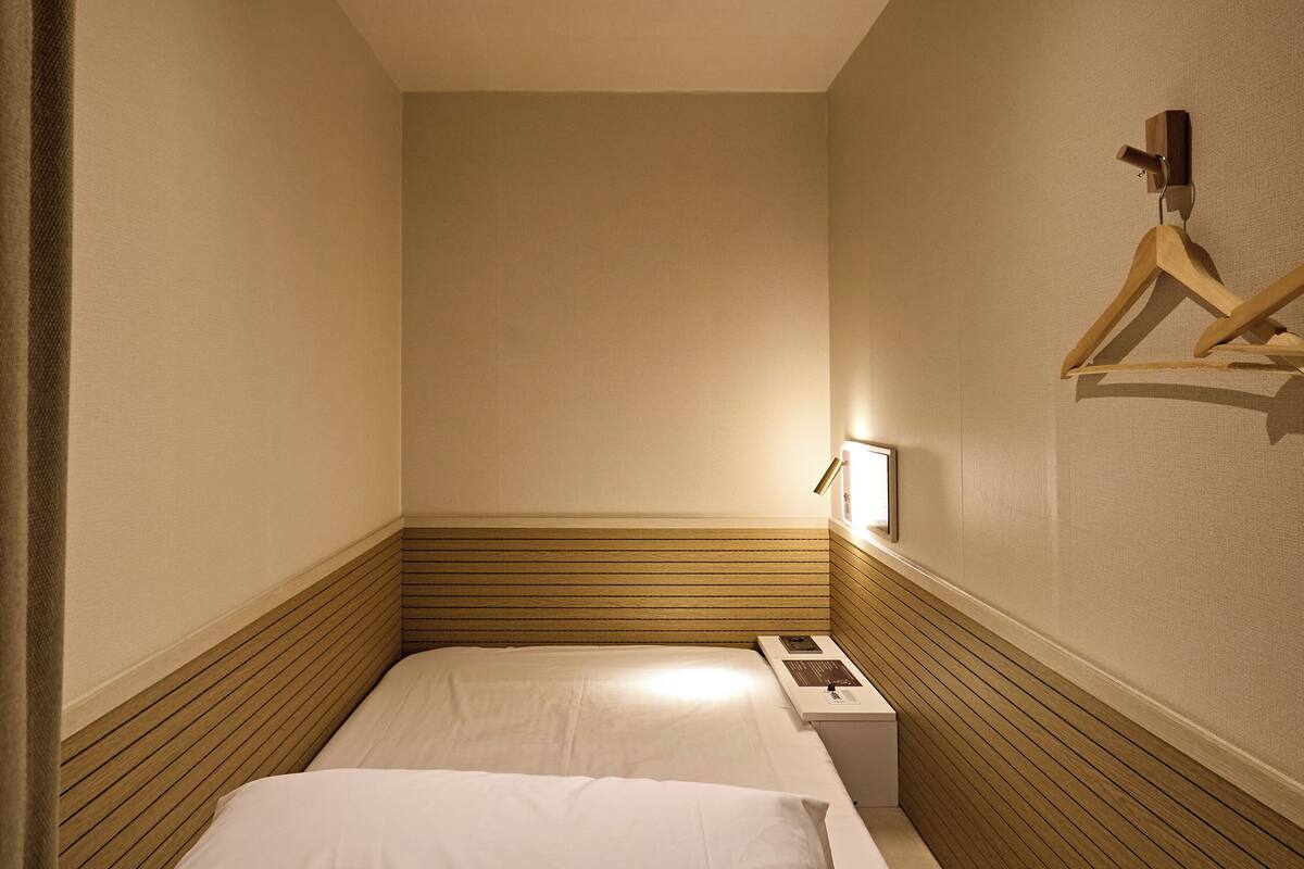 A narrow hotel room with a single bed, a bedside table, and a wall-mounted hanger.