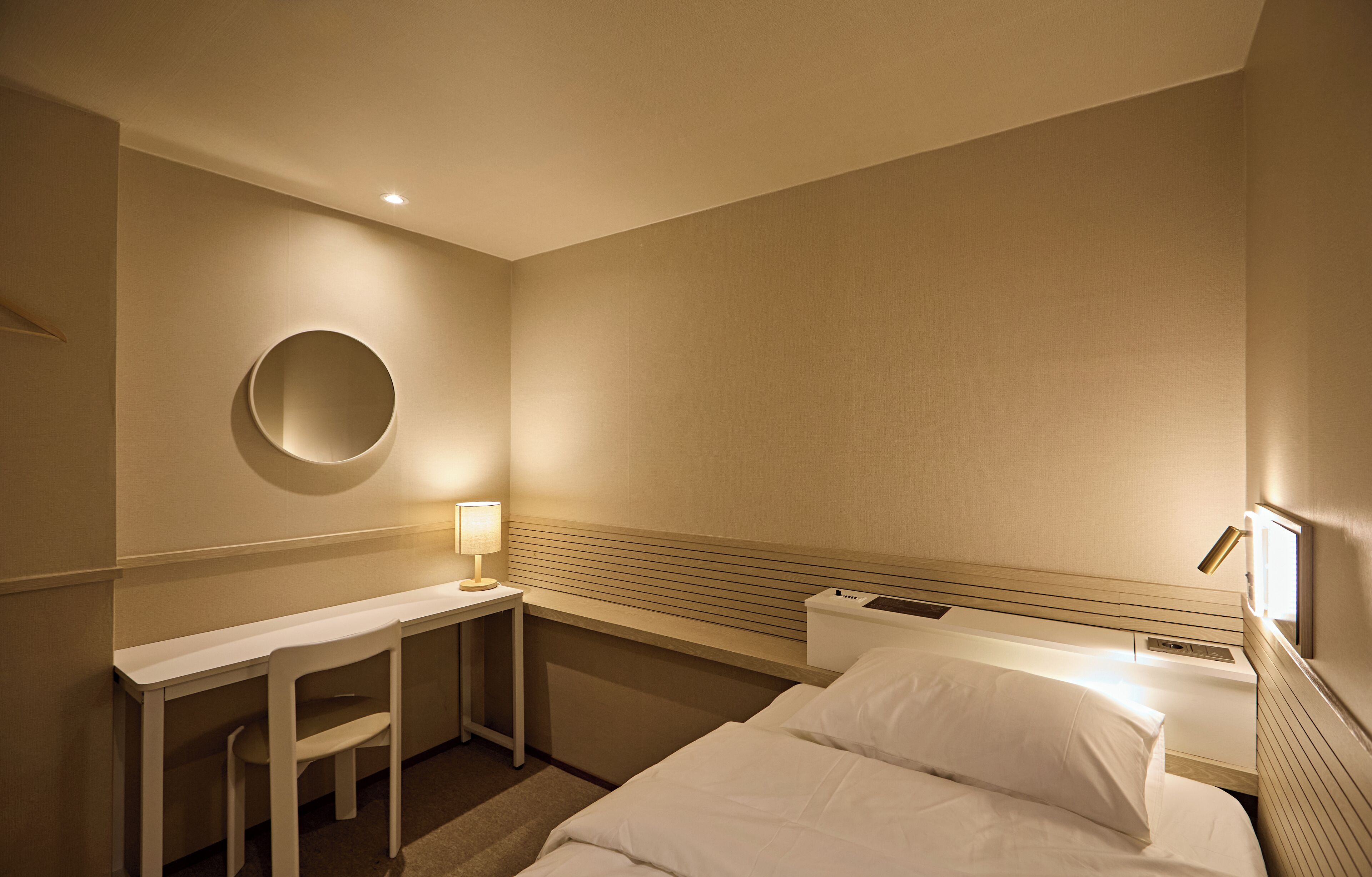A hotel room with a bed, a desk, a chair, a round mirror, and a nightstand with a lamp.