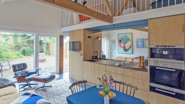 A modern kitchen with a built-in oven, a dining area with a blue tablecloth, and a view of the garden through the open door. - Sparrows Nest - UK50109 (Sparrow Green, near Dereham)
